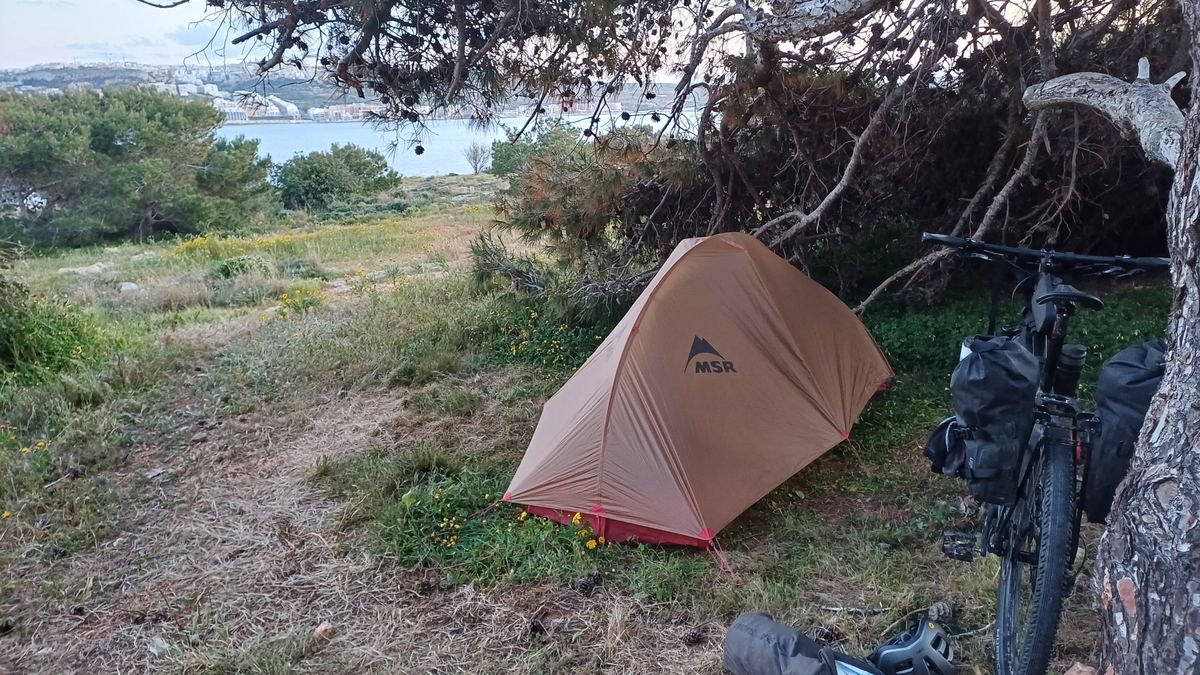 Zelt unter einer Kiefer mit Blick auf eine Bucht auf Malta, daneben ein beladenes Fahrrad
